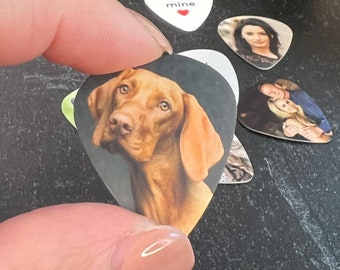 May include: A collection of guitar picks featuring custom images. One pick displays a brown dog, while others showcase portraits and the text "be mine" with a heart. The picks are in various shapes and sizes.