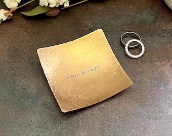 May include: A square, gold-toned metal trinket dish with a hammered texture and the text "Love Always" stamped in the center. Two silver-toned rings, one plain and one with a beaded edge, are positioned next to the dish on the surface.