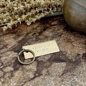 May include: A rectangular, hammered metal keychain with the phrase "I LOVE YOU MOST" and a heart symbol. The keychain has a bronze-colored key ring. The background features dried plants and a wooden vase.