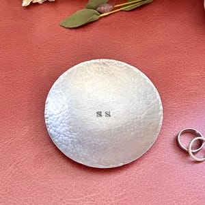 May include: A hammered silver-colored ring dish with a textured surface. Two silver rings are next to the dish. The dish has a series of vertical lines etched into the center. The background is a reddish-pink color.