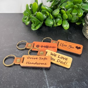 May include: Four wooden keychains with different messages engraved on them. The keychains are brown, red, and natural wood. The messages are "Drive Safe Handsome", "We Love You Dad!", "I Love You", and a sound wave.