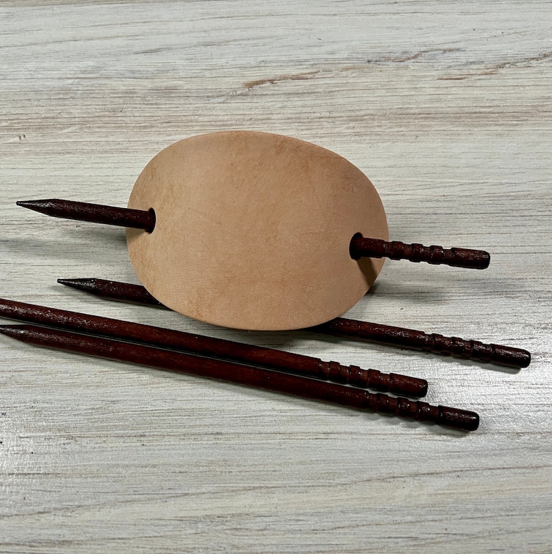 May include: A light brown leather hair clip with three dark brown wooden hair sticks. The hair sticks have a textured surface.