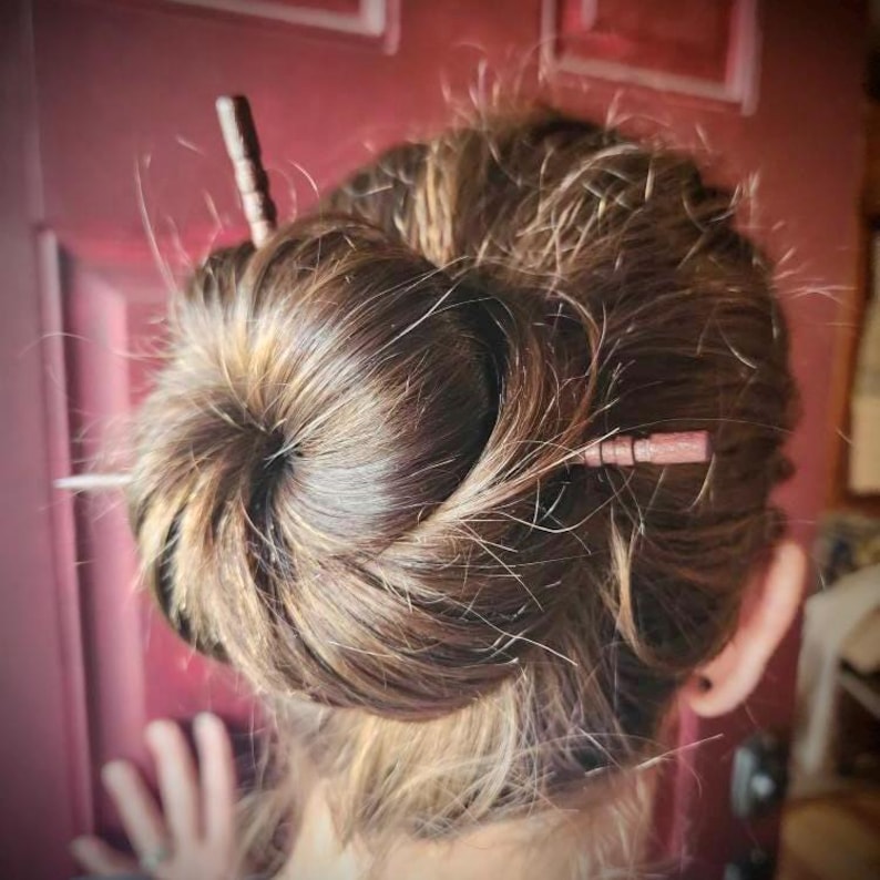 May include: A woman with long brown hair styled in a bun with two wooden hair sticks. The hair sticks are brown with a dark brown finish.