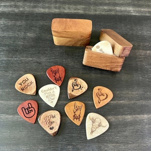 May include: A collection of guitar picks with various designs and text, including a rock and roll hand gesture, a treble clef heart, and the words "You Rock" and "I Pick You." The picks are displayed with two small wooden boxes.