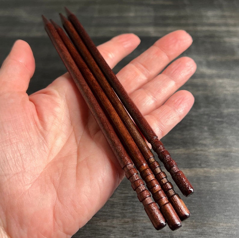 May include: Four wooden skewers with a dark red stain and a textured pattern. The skewers are held in a hand.