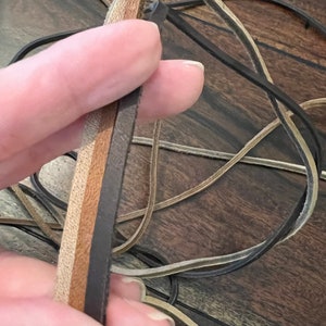 May include: Close-up of leather strips in various shades of brown and tan. The strips are held in a hand, with other leather cords visible in the background. The leather appears to be of different textures and thicknesses, suitable for crafting or jewelry making.