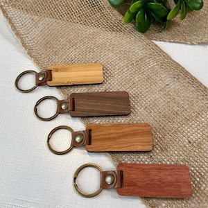 May include: Four wooden keychains with brass key rings and leather straps. The keychains are made of different types of wood, including bamboo, walnut, cherry, and redwood. The keychains are rectangular and have a natural wood grain.