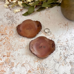 May include: Two brown, decorative ring dishes with ornate edges. One dish has the text "I STILL DO", the other has tally marks. Two silver rings are also present. The dishes are on a patterned surface.