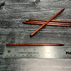 May include: Five wooden skewers with a brown finish and a decorative pattern. The skewers are measured against a stainless steel ruler, showing they are approximately 6 inches long.
