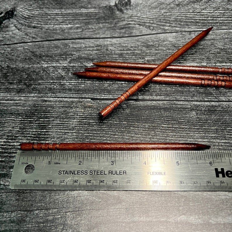 May include: Five wooden skewers with a brown finish and a decorative pattern. The skewers are measured against a stainless steel ruler, showing they are approximately 6 inches long.