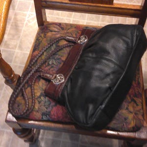 K&ouml;nnte beinhalten: Eine schwarze Ledertasche mit dunkelbraunem Lederbesatz und geflochtenen Griffen. Die Tasche ist mit silberfarbenen, herzf&ouml;rmigen Metallbeschl&auml;gen an der Basis jedes Griffs akzentuiert, die dem strukturierten Design ein dekoratives Detail verleihen.