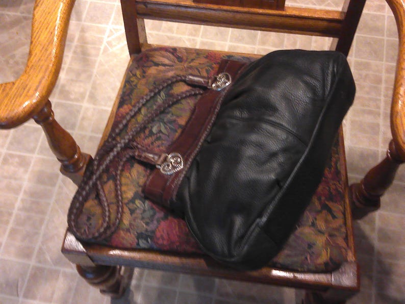 K&ouml;nnte beinhalten: Eine schwarze Ledertasche mit dunkelbraunem Lederbesatz und geflochtenen Griffen. Die Tasche ist mit silberfarbenen, herzf&ouml;rmigen Metallbeschl&auml;gen an der Basis jedes Griffs akzentuiert, die dem strukturierten Design ein dekoratives Detail verleihen.