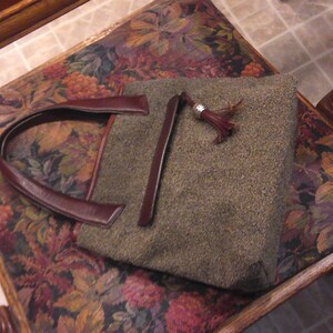 May include: A rectangular handbag featuring a textured olive-green wool-like fabric body, dark burgundy leather handles, and a matching leather zipper pull adorned with a silver-toned metal tassel cap. The bag has a structured, flat-bottomed design.