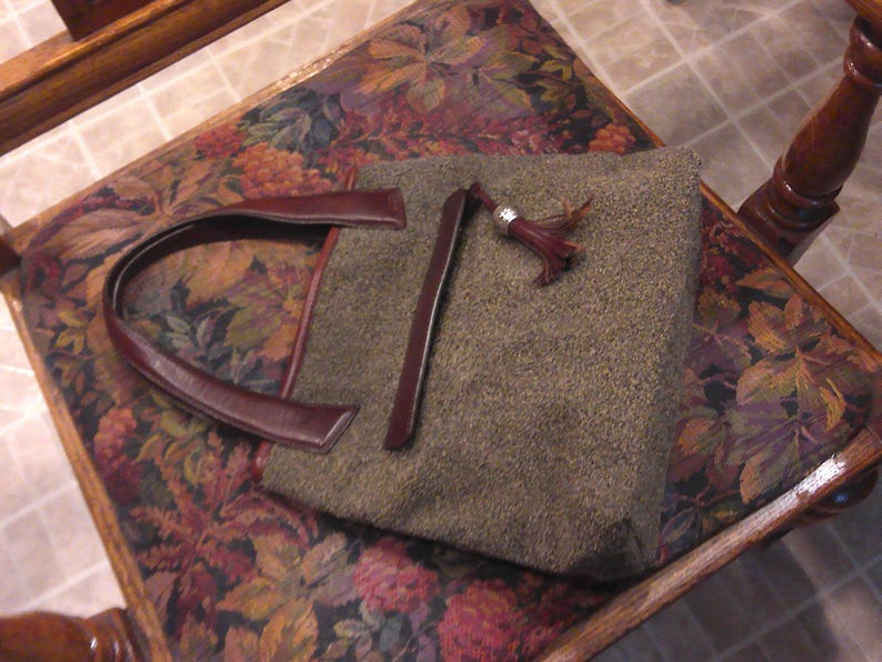 May include: A rectangular handbag featuring a textured olive-green wool-like fabric body, dark burgundy leather handles, and a matching leather zipper pull adorned with a silver-toned metal tassel cap. The bag has a structured, flat-bottomed design.
