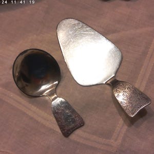 May include: Two silver spoons with floral designs on the handles. The spoon on the left is a small serving spoon, and the spoon on the right is a cake server.
