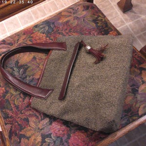 May include: A structured handbag featuring a textured, olive-green wool-like fabric body. It includes dark burgundy leather handles and a matching leather trim along the top edge. A decorative leather tassel with a silver-toned metal cap is attached.