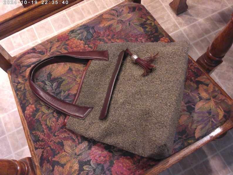 May include: A structured handbag featuring a textured, olive-green wool-like fabric body. It includes dark burgundy leather handles and a matching leather trim along the top edge. A decorative leather tassel with a silver-toned metal cap is attached.