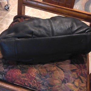 K&ouml;nnte beinhalten: Eine schwarze Handtasche aus Leder mit genarbter Textur und einer abgerundeten, weichen Silhouette. Die Tasche verf&uuml;gt &uuml;ber einen breiten, flachen Boden und einen geflochtenen braunen Ledergriff, der oben teilweise sichtbar ist und einen Kontrast bildet.
