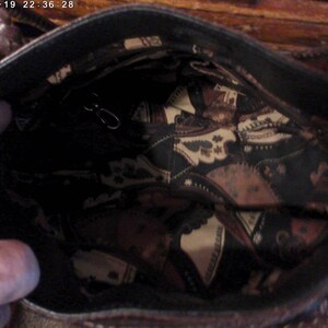 May include: The interior of a dark brown handbag featuring a black lining with a repeating cream and brown paisley-style pattern. The bag includes a small metal clasp attached to a thin strap inside the main compartment and braided handles.