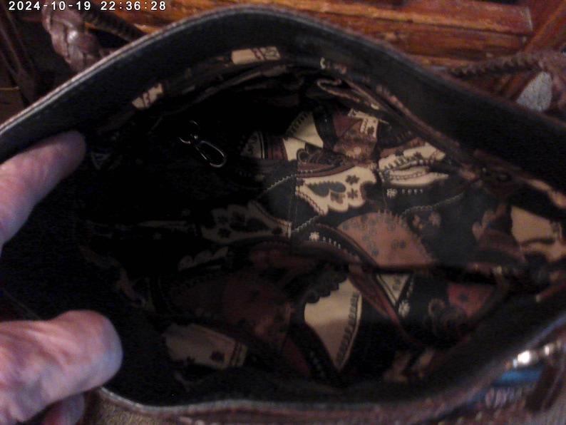 May include: The interior of a dark brown handbag featuring a black lining with a repeating cream and brown paisley-style pattern. The bag includes a small metal clasp attached to a thin strap inside the main compartment and braided handles.