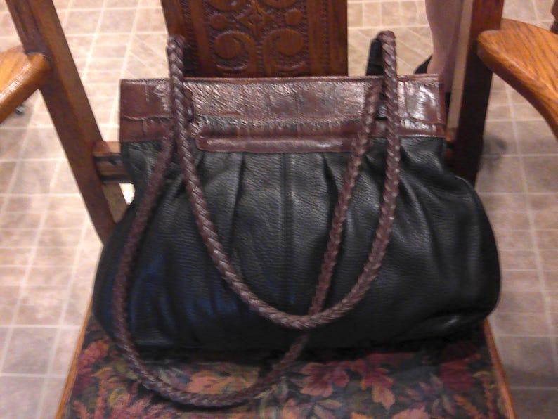 K&ouml;nnte beinhalten: Eine schwarze Ledertasche mit einem dunkelbraunen, krokodilgepr&auml;gten Lederbesatz am oberen Rand und zwei geflochtenen dunkelbraunen Lederriemen. Der Hauptteil der Tasche hat ein gerafftes, gefaltetes Design mit einer weichen, strukturierten Oberfl&auml;che.