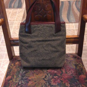 May include: A rectangular handbag featuring a textured, olive-green fabric body and dark brown leather handles. The bag has a simple, structured design with a flat base and a top opening, showcasing a contrast between the fuzzy material and leather.