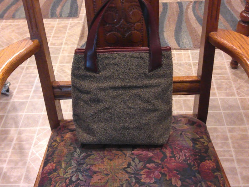 May include: A rectangular handbag featuring a textured, olive-green fabric body and dark brown leather handles. The bag has a simple, structured design with a flat base and a top opening, showcasing a contrast between the fuzzy material and leather.