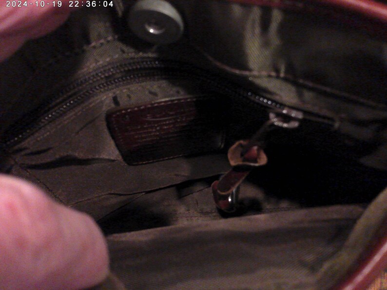 May include: The interior of a dark brown leather bag featuring a dark olive green fabric lining, a zippered pocket with a leather brand patch, and a magnetic snap closure at the top. The bag includes a small leather zipper pull tab.