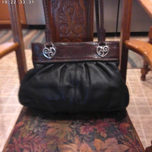 K&ouml;nnte beinhalten: Eine schwarze Ledertasche mit gerafftem, gefaltetem K&ouml;rper und einem glatten, dunkelbraunen Leder-Oberteil. Die Tasche verf&uuml;gt &uuml;ber zwei geflochtene Lederriemen, die mit silberfarbenen Metallbeschl&auml;gen in Form von durchbrochenen Herzen befestigt sind.