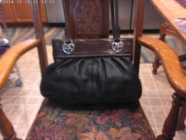 K&ouml;nnte beinhalten: Eine schwarze Ledertasche mit gerafftem, gefaltetem K&ouml;rper und einem glatten, dunkelbraunen Leder-Oberteil. Die Tasche verf&uuml;gt &uuml;ber zwei geflochtene Lederriemen, die mit silberfarbenen Metallbeschl&auml;gen in Form von durchbrochenen Herzen befestigt sind.