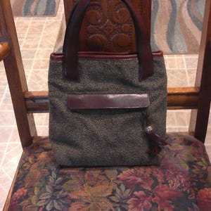 May include: A structured handbag featuring a textured, dark gray tweed-like fabric body with dark brown leather trim, two matching leather handles, a horizontal leather flap pocket on the front, and a small leather tassel with a silver-toned accent.