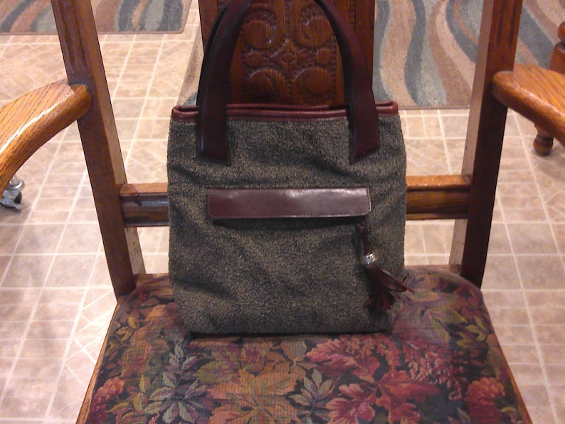 May include: A structured handbag featuring a textured, dark gray tweed-like fabric body with dark brown leather trim, two matching leather handles, a horizontal leather flap pocket on the front, and a small leather tassel with a silver-toned accent.