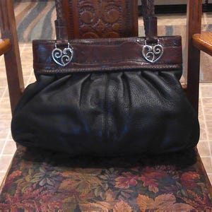 K&ouml;nnte beinhalten: Eine schwarze Ledertasche mit gerafftem, gefaltetem K&ouml;rper und einem glatten, dunkelbraunen Leder-Oberteil. Die Tasche verf&uuml;gt &uuml;ber zwei geflochtene Ledergriffe, die mit silberfarbenen Metallbeschl&auml;gen in Form von verzierten, geschwungenen Herzen befestigt sind.