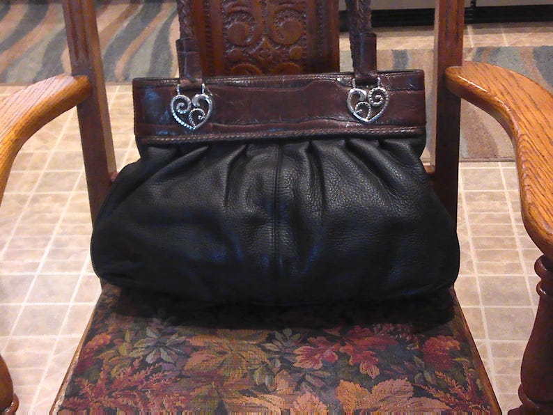 K&ouml;nnte beinhalten: Eine schwarze Ledertasche mit gerafftem, gefaltetem K&ouml;rper und einem glatten, dunkelbraunen Leder-Oberteil. Die Tasche verf&uuml;gt &uuml;ber zwei geflochtene Ledergriffe, die mit silberfarbenen Metallbeschl&auml;gen in Form von verzierten, geschwungenen Herzen befestigt sind.