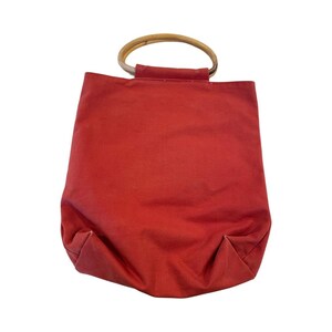 May include: A red canvas tote bag with a wooden handle. The bag has a simple design and is perfect for carrying everyday essentials.