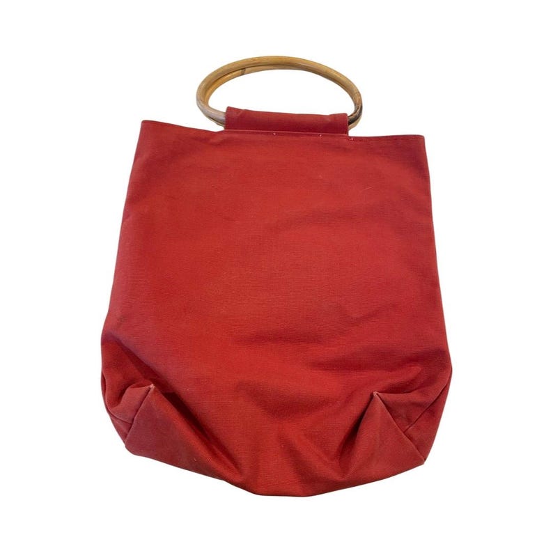 May include: A red canvas tote bag with a wooden handle. The bag has a simple design and is perfect for carrying everyday essentials.