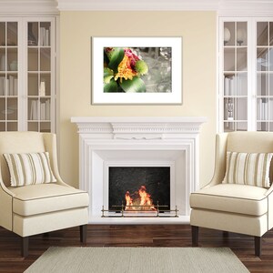May include: A white fireplace with a fire burning inside. Two beige armchairs with striped pillows are placed in front of the fireplace. A framed print of a colorful flower arrangement hangs above the fireplace.