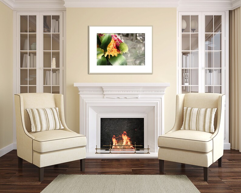 May include: A white fireplace with a fire burning inside. Two beige armchairs with striped pillows are placed in front of the fireplace. A framed print of a colorful flower arrangement hangs above the fireplace.