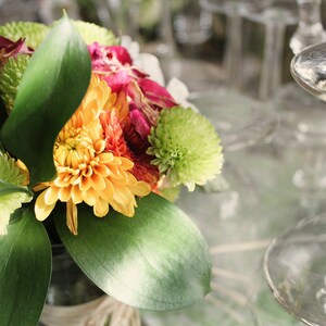 May include: A bouquet of flowers with yellow, orange, pink, and green blooms. The flowers are arranged in a clear glass vase with green leaves.