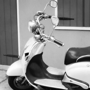 May include: A white and silver scooter with a black seat. The scooter has a chrome handlebar with a rearview mirror on each side. The scooter is parked in front of a building with a wooden door and a window.