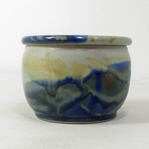 Op de afbeelding: Een keramische botervloot met een witte basis en blauwe en gele glazuur. De vloot heeft een deksel.