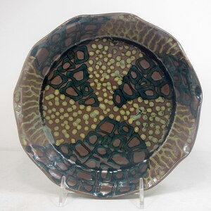 Puede incluir: Un plato para tartas de cerámica marrón con un borde festoneado. El plato está decorado con un patrón de puntos y formas verdes, marrones y amarillos.