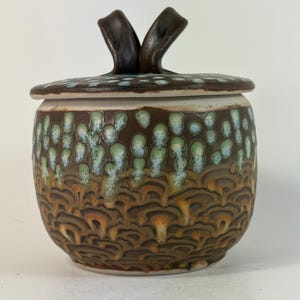 May include: A ceramic lidded jar with a dark brown base featuring a textured, scalloped pattern. The upper portion displays a glaze of light green and white spots. The lid has a dark brown, curved handle extending from the center.