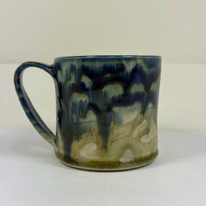May include: A ceramic mug with a curved handle. The mug features a glaze with a blend of dark blue, green, and tan colors. The glaze has a dripped effect, creating a unique and artistic appearance. The mug is suitable for hot or cold beverages.