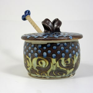 May include: A brown and white ceramic honey pot with a blue and brown lid and a wooden dipper with a blue handle. The pot has a swirl pattern and blue dots.