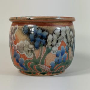 May include: A round ceramic jar with a lid, featuring a hand-painted floral pattern in shades of blue, white, and green against a warm orange and tan background. The vessel has a smooth, glossy finish and a slightly tapered shape with a flat rim.