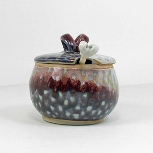 May include: A ceramic honey pot with a lid and a small spoon. The pot is decorated with a purple, blue, and white glaze. The lid has a decorative bow.