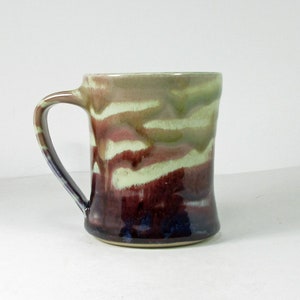 May include: A ceramic mug with a white, green, and purple glaze. The mug has a wide, rounded base and a handle.