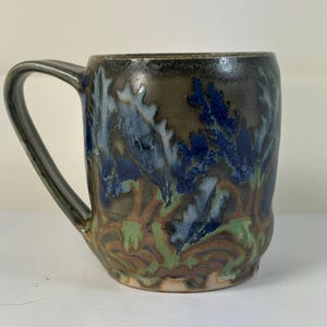 May include: A handmade ceramic mug with a dark handle and a unique glaze. The mug features a blend of colors, including blue, green, brown, and black, creating a textured, organic pattern. The mug is designed for hot beverages.