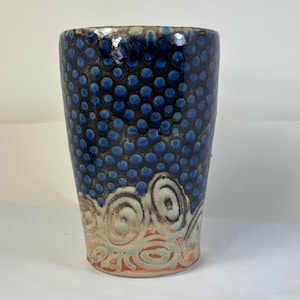 May include: A handmade ceramic tumbler with a textured surface. The upper portion features a dark blue background with raised blue dots. The lower section has a white, orange, and green spiral design. The rim is a light brown color.